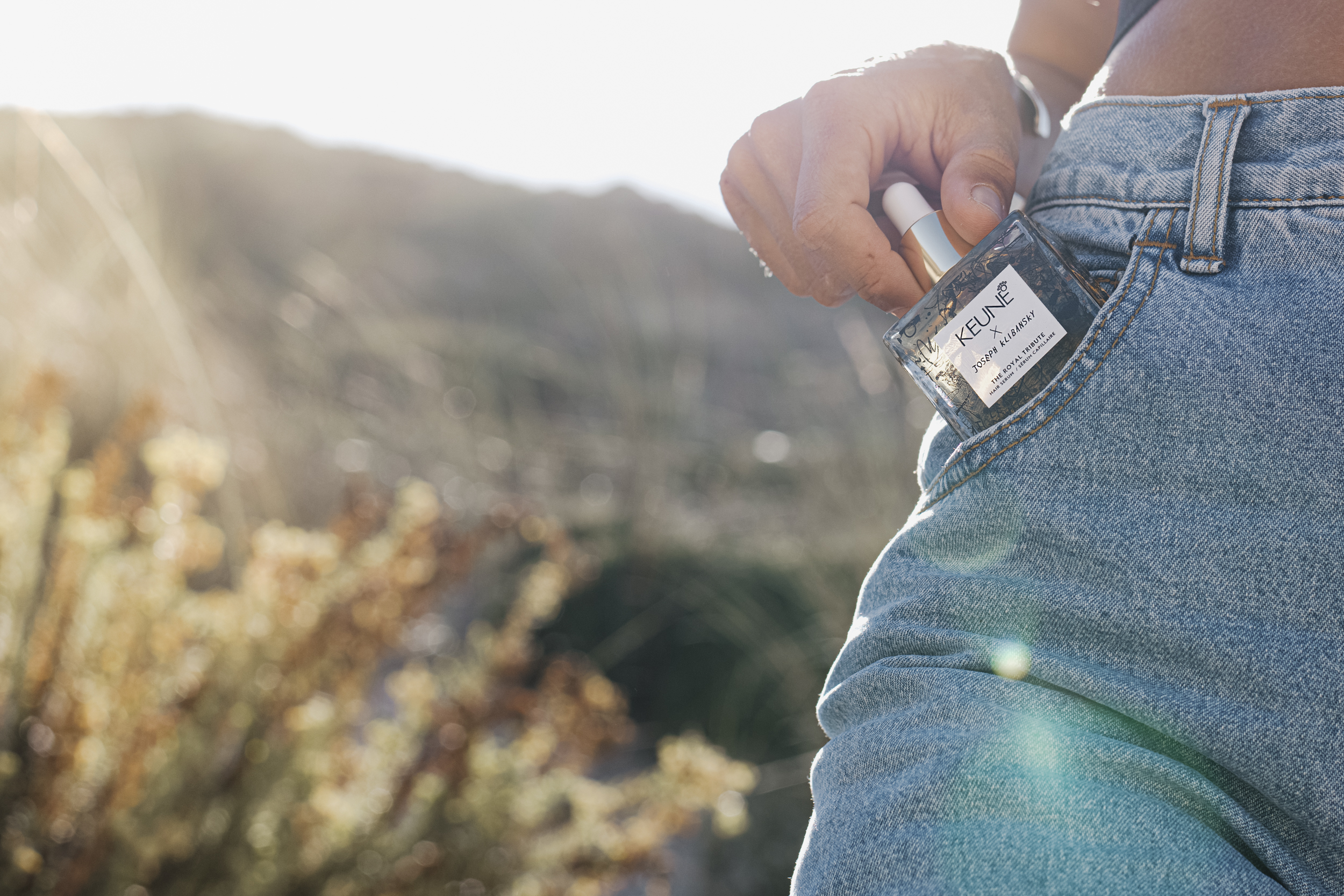 Eine Frauenhand zieht aus einer Jeans im Gegenlicht eine Glasflasche Pflegeöl aus der Jeans