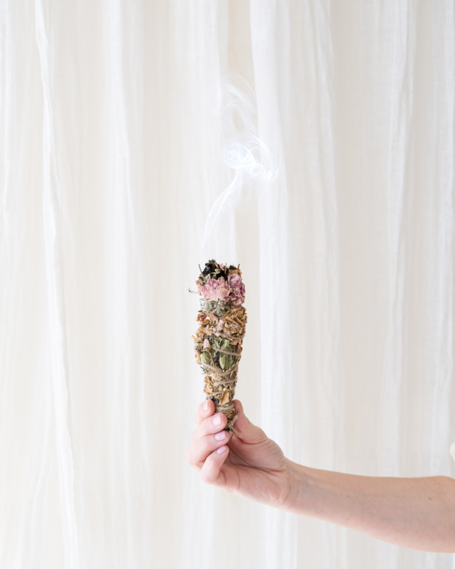 Frauenhand vor einem hellen Vorhang. Hält Weihrauchgebinde zum ausräuchern. Rauch steigt in Kräuseln auf.