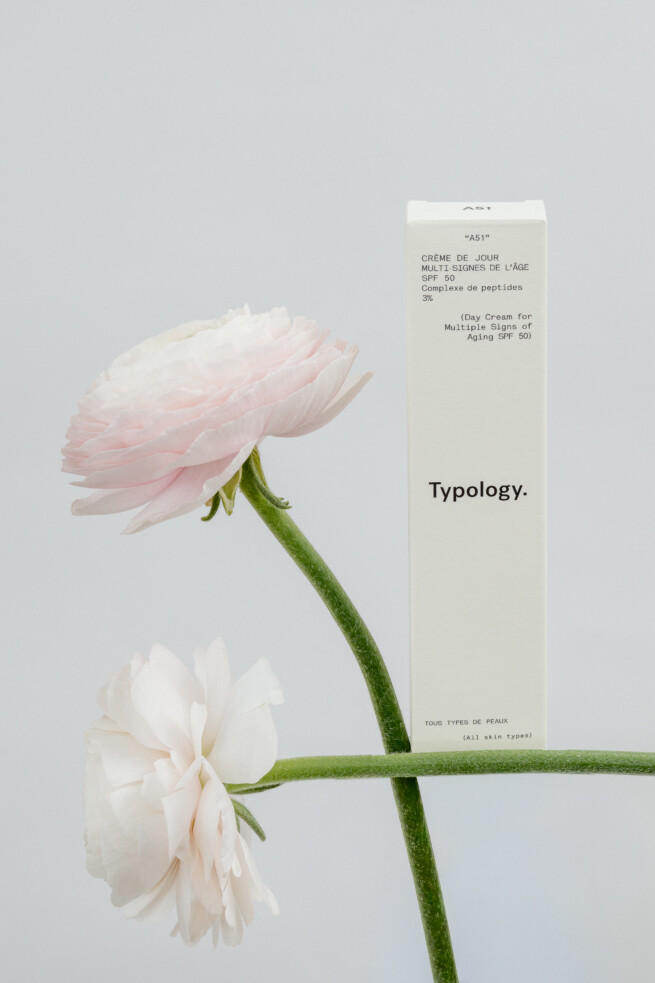 Typology Tagescreme SPF 50 neben zarten rosafarbenen Blüten, fotografiert in einer minimalistischen und natürlichen Beauty-Inszenierung.